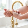 Kobiece dłonie wyciskające wodę z mokrej, beżowej ściereczki z mikrofibry nad złotą baterią zlewozmywakową.