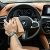 Męska dłoń z zegarkiem i tatuażem przecierająca czarną skórzaną kierownicę w samochodzie marki BMW za pomocą beżowej ściereczki z mikrofibry.