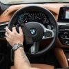 CMęska dłoń z zegarkiem i tatuażem przecierająca czarną skórzaną kierownicę w samochodzie marki BMW za pomocą beżowej ściereczki z mikrofibry.