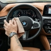 Męska dłoń z zegarkiem i tatuażem przecierająca czarną skórzaną kierownicę w samochodzie marki BMW za pomocą beżowej ściereczki z mikrofibry.