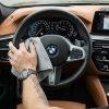 Męska dłoń z tatuażem i zegarkiem przecierająca skórzaną kierownicę w samochodzie marki BMW za pomocą szarej ściereczki z mikrofibry