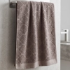 Szarobrązowy (taupe) ręcznik bawełniany frotte Fiamo (50x100) w modnym, eleganckim stylu, zawieszony na stalowym relingu na tle jasnych płytek.