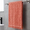 Koralowy ręcznik bawełniany frotte Lana (50x100) z frędzlami w modnym stylu boho, zawieszony na stalowym relingu na tle szarych płytek.