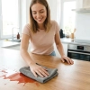 Uśmiechnięta kobieta wycierająca rozlany płyn z drewnianego blatu kuchennego szarą magiczną ściereczką z mikrofibry