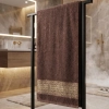 Ciemnobrązowy chłonny ręcznik PERLA ze złotym akcentem zawieszony na czarnym stojaku w łazience.