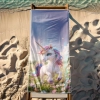Szybkoschnący ręcznik plażowy z tęczowym jednorożcem rozłożony na drewnianym leżaku stojącym na piasku nad brzegiem morza.