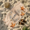 Wielki ręcznik plażowy Serena z kotwicą z muszli i rozgwiazdami, leżący na piasku obok słomkowego kapelusza i brązowych klapków
