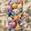 Duży ręcznik plażowy XXL z mikrofibry ze wzorem wielokolorowych muszli i rozgwiazdy, rozłożony na piasku obok kapelusza