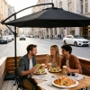 Trzy uśmiechnięte osoby jedzące śniadanie i pijące kawę przy drewnianym stole na zewnętrznym patio, osłonięte eleganckim, czarnym parasolem ogrodowym.