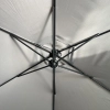 Złożony parasol ogrodowy na wysięgniku Sombra w kolorze szarym z dociążoną podstawą krzyżową, stojący na tarasie obok kwitnących krzewów w ogrodzie.