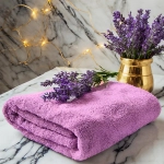Puszysty ręcznik frotte do twarzy 50x100 w kolorze lawendowym, ułożony w kostkę na jasnym marmurze z kwiatami.