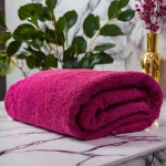 Puszysty ręcznik frotte do twarzy 50x100 w kolorze biskupim, ułożony na jasnym marmurze przy zielonych liściach.