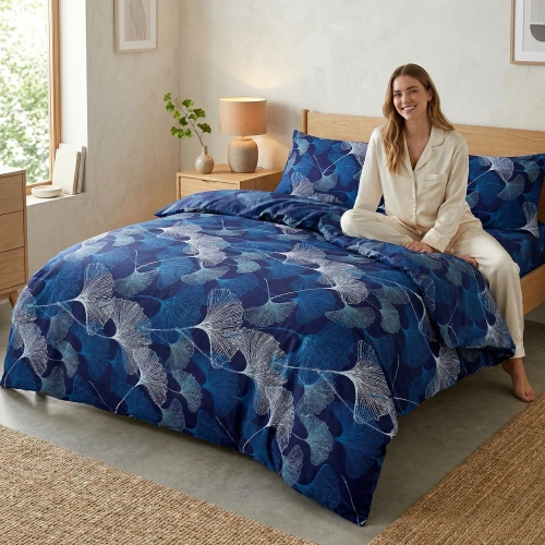 Uśmiechnięta kobieta w jasnej piżamie siedzi na łóżku zaścielonym granatową pościelą w jasne liście miłorzębu marki J&W.