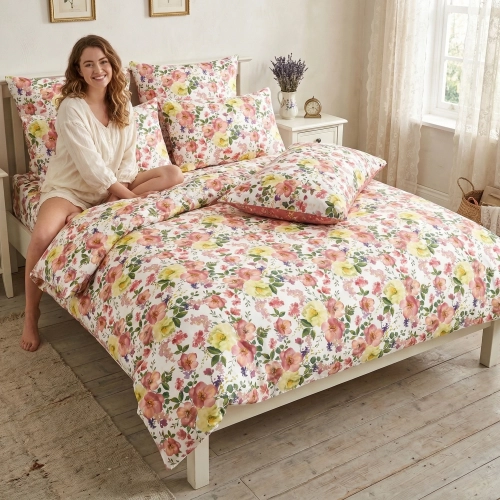 Uśmiechnięta kobieta w jasnej piżamie siedzi na łóżku z piękną, kwiatową pościelą z bawełny premium J&W.