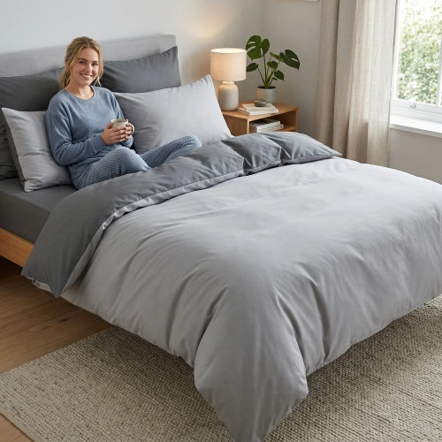 Uśmiechnięta kobieta relaksuje się w łóżku z dwustronną, jasnoszaro-grafitową pościelą J&W.