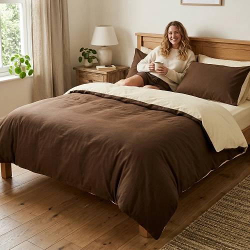 Uśmiechnięta kobieta relaksuje się w łóżku z dwustronną, brązowo-beżową pościelą J&W.