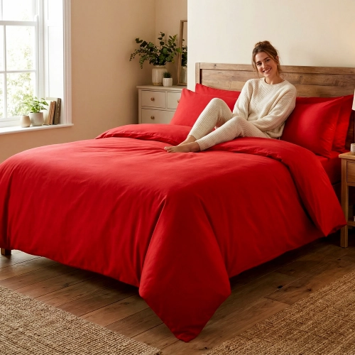 Uśmiechnięta kobieta relaksuje się w łóżku z jednokolorową, czerwoną pościelą z bawełny premium J&W.