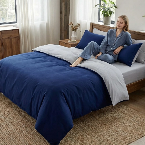 Uśmiechnięta kobieta relaksuje się w łóżku z dwustronną, granatowo-jasnoszarą pościelą J&W.