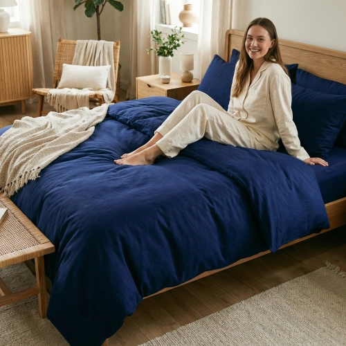Uśmiechnięta kobieta relaksuje się siedząc na łóżku z jednokolorową, granatową pościelą z bawełny premium J&W.
