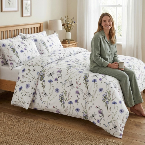 Uśmiechnięta kobieta w zielonej piżamie siedzi na łóżku z białą pościelą z bawełny premium J&W w polne chabry i ważki.