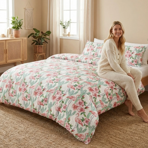 Uśmiechnięta kobieta w jasnym swetrze siedzi na łóżku z uroczą pościelą z bawełny premium J&W w różowe flamingi i kaktus