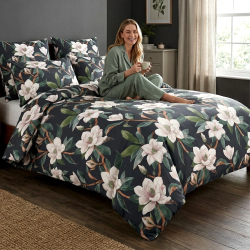 Uśmiechnięta kobieta w szałwiowej piżamie siedzi z kubkiem na łóżku z elegancką, ciemną pościelą z bawełny premium J&W w