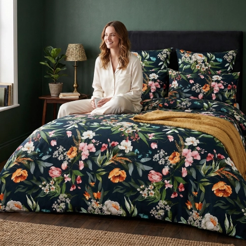 Uśmiechnięta kobieta w jasnej piżamie siedzi na łóżku zaścielonym ciemną pościelą z bawełny premium J&W w bogaty wzór kw