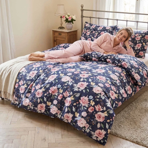 Kobieta uśmiecha się leżąc na łóżku z granatową pościelą z bawełny premium J&W, ubraną we wzór Nocna Róża.