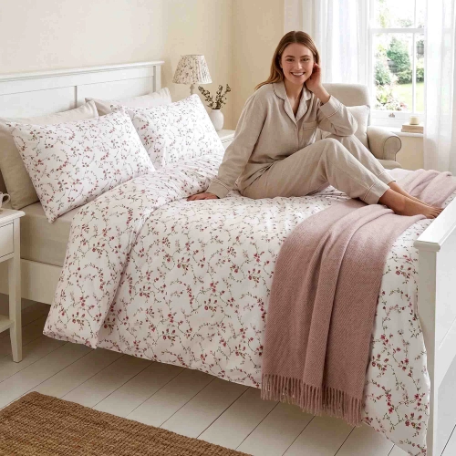 Uśmiechnięta kobieta w jasnej piżamie relaksuje się na łóżku w sypialni, zaścielonym w pościel "Pudrowy Ogród" J&W w róż