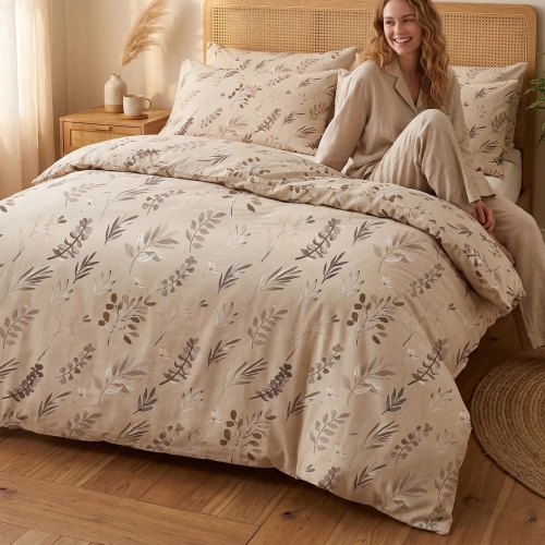 Kobieta w beżowej piżamie siedzi uśmiechnięta na łóżku ubranym w bawełnianą pościel J&W we wzór gałązek w barwach ziemi.