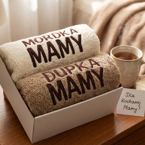 Zestaw dwóch ręczników z haftem Mordka Mamy i Dupka Mamy w eleganckim pudełku na drewnianym stole obok kubka herbaty.