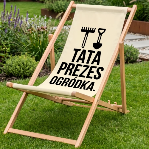 Drewniany leżak ogrodowy w kolorze ecru na zielonym trawniku z nadrukiem TATA PREZES OGRÓDKA oraz grabiami i łopatą, tło