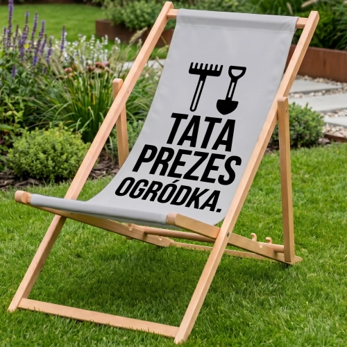 Drewniany leżak ogrodowy w kolorze jasnoszarym na zielonym trawniku z nadrukiem TATA PREZES OGRÓDKA oraz grabiami i łopa