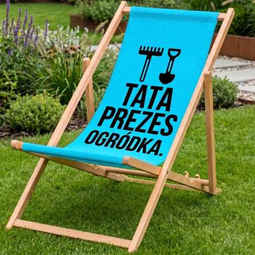 Drewniany leżak ogrodowy w kolorze niebieskim na zielonym trawniku z nadrukiem TATA PREZES OGRÓDKA oraz grabiami i łopat