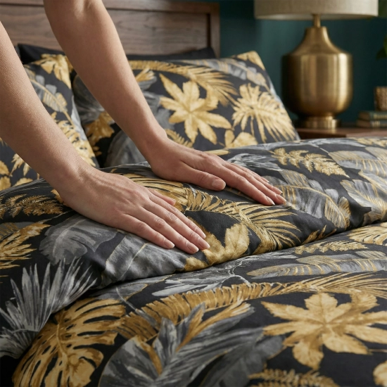 Zbliżenie na dłonie gładzące miękki, bawełniany materiał pościeli z wyrazistym, tropikalnym motywem złotych liści.