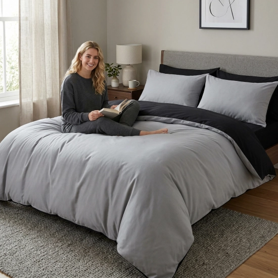 Uśmiechnięta kobieta siedzi z książką na łóżku z dwustronną, jasnoszaro-czarną pościelą J&W.