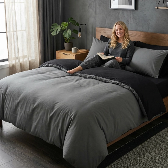 Uśmiechnięta kobieta czyta książkę w łóżku z dwustronną, ciemnoszaro-czarną pościelą J&W.