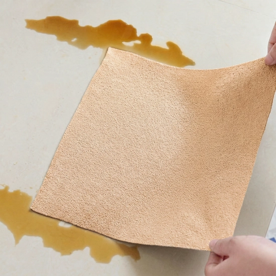 Dwie dłonie trzymające beżową ściereczkę z mikrofibry, która wchłania rozlaną brązową ciecz z jasnych płytek podłogowych.