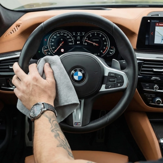 Męska dłoń z tatuażem i zegarkiem przecierająca skórzaną kierownicę w samochodzie marki BMW za pomocą szarej ściereczki z mikrofibry