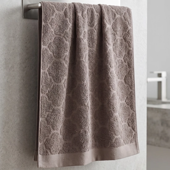 Szarobrązowy (taupe) ręcznik bawełniany frotte Fiamo (50x100) w modnym, eleganckim stylu, zawieszony na stalowym relingu na tle jasnych płytek.