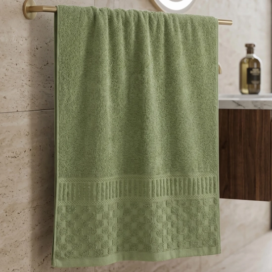 Szałwiowo-zielony ręcznik bawełniany zawieszony na złotym wieszaku na tle beżowej ściany z płytek