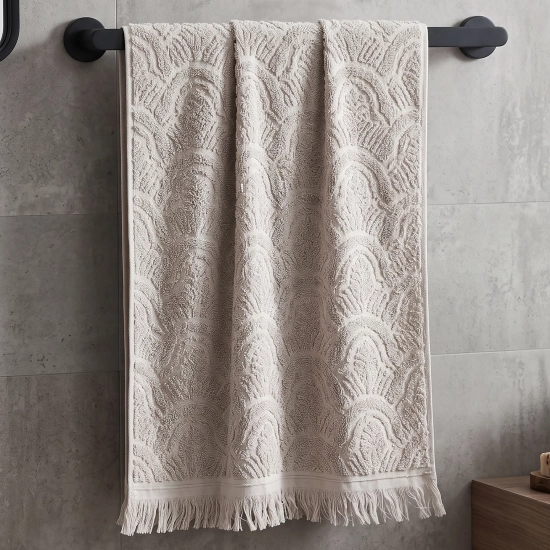 Jasnoszary, klasyczny ręcznik bawełniany Lana 50x100 w żakardowy wzór w fale, wykończony na dole eleganckimi frędzlami, wiszący na relingu ściennym.