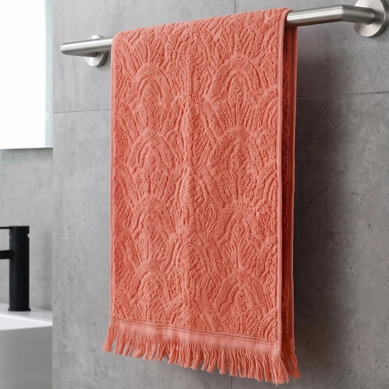 Koralowy ręcznik bawełniany frotte Lana (50x100) z frędzlami w modnym stylu boho, zawieszony na stalowym relingu na tle szarych płytek.