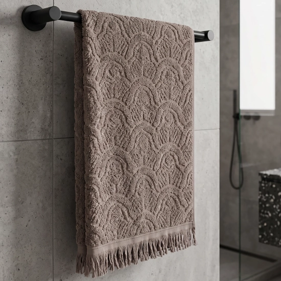 Brązowy (taupe) ręcznik do rąk i twarzy z kolekcji Lana 50x100 w elegancki wzór żakardowych łuków z frędzlami, powieszony na czarnym wieszaku.