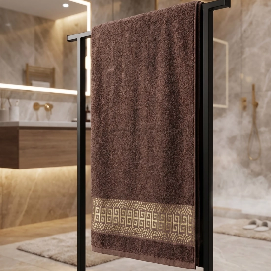 Ciemnobrązowy chłonny ręcznik PERLA ze złotym akcentem zawieszony na czarnym stojaku w łazience.