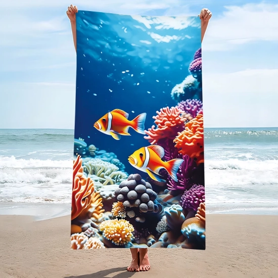 Pionowa prezentacja dużego ręcznika plażowego we wzór oceanu i uroczych rybek, trzymanego przez osobę stojącą na piaszczystej plaży