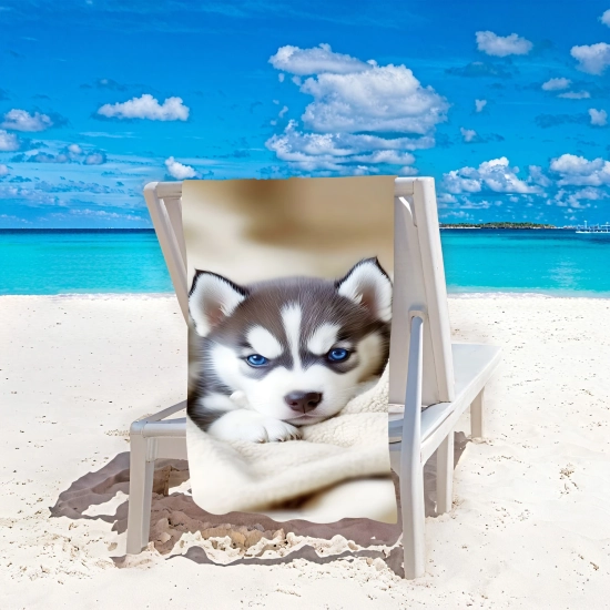 Duży ręcznik plażowy XXL z mikrofibry w małego pieska husky przewieszony przez biały leżak stojący na białym piasku na tle oceanu