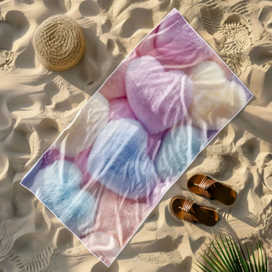 Lekki ręcznik plażowy z motywem futrzanych serc rozłożony na piasku obok słomkowego kapelusza i brązowych klapek