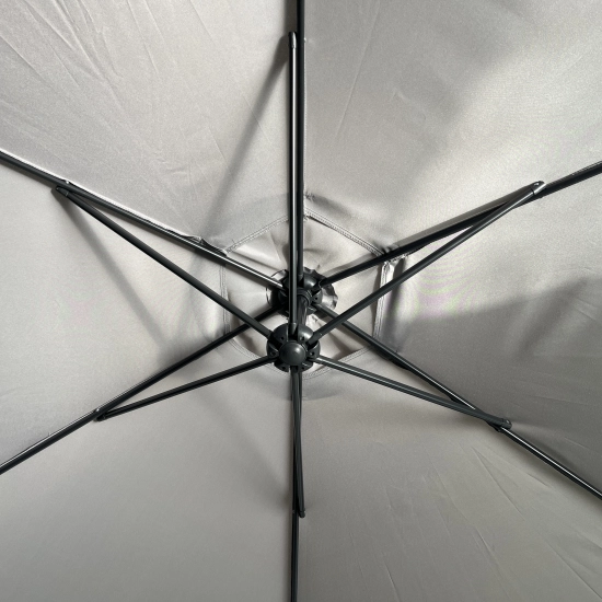 Złożony parasol ogrodowy na wysięgniku Sombra w kolorze szarym z dociążoną podstawą krzyżową, stojący na tarasie obok kwitnących krzewów w ogrodzie.