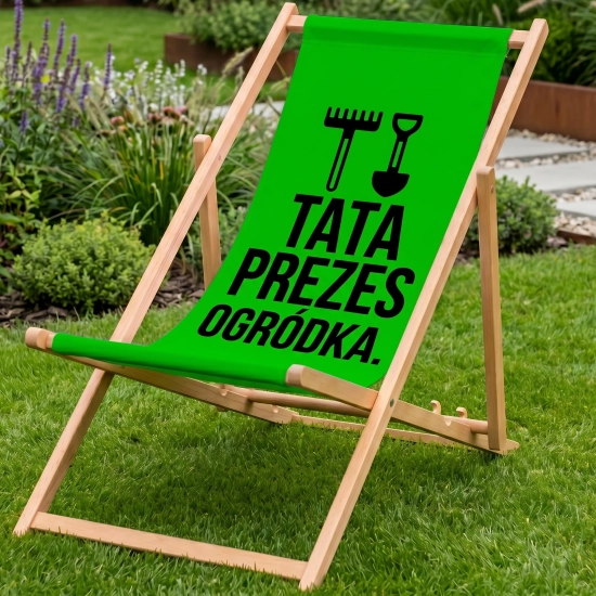 Drewniany leżak ogrodowy w kolorze zielonym na zielonym trawniku z nadrukiem TATA PREZES OGRÓDKA oraz grabiami i łopatą,